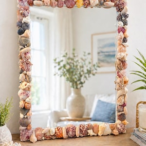May include: Square mirror with a frame entirely decorated with various seashells in shades of white, pink, orange, and brown. The mirror reflects a room with a vase of flowers and a window. The mirror is a decorative coastal accent.