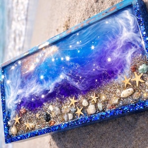 May include: A rectangular resin art piece with a beach theme. The artwork features a blue glitter frame, a swirling blue and purple ocean design, small starfish, and pebbles. The piece is resting on sand.