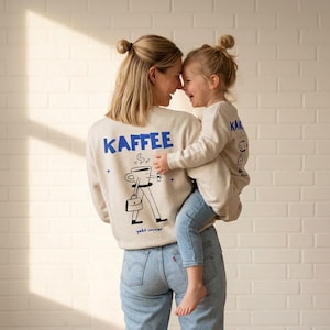 Può includere: Felpe beige con testo blu e grafica a tema caffè. La felpa per adulti dice "KAFFEE" e presenta un personaggio a forma di tazza di caffè che porta una valigetta. La felpa per bambini ha un design simile. Entrambi indossano jeans blu.