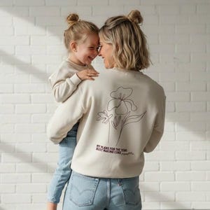 Può includere: Una felpa beige con un disegno floreale e la scritta "MY PLANS FOR THE YEAR: BUSY MAKING CORE MEMORIES." Una donna tiene in braccio una bambina, entrambe sorridenti. Indossano felpe chiare e jeans blu.