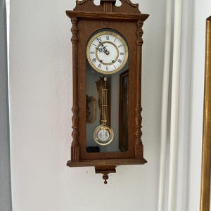 Könnte beinhalten: Eine Wanduhr aus Holz mit rechteckigem Rahmen. Das Zifferblatt ist weiß mit schwarzen römischen Ziffern und goldenen Zeigern. Ein Pendel schwingt im verglasten Bereich. Die Uhr ist an einer weißen Wand befestigt.