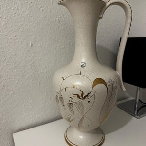 Könnte beinhalten: Cremefarbener Keramikkrug mit schmalem Hals und gebogenem Henkel. Der Krug hat goldfarbene Blumendesigns und einen Craquelé-Effekt. Ein kleines Emblem ist oben sichtbar. Der Sockel hat einen goldenen Rand.