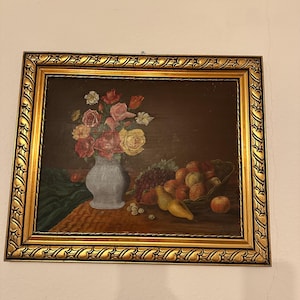 Könnte beinhalten: Gerahmtes Stillleben-Gemälde mit einer Vase mit Rosen und anderen Blumen sowie einem Obstkorb. Das Gemälde hat einen dunklen Hintergrund und ist in einem goldenen, verzierten Rahmen gerahmt. Das Obst umfasst Trauben, Äpfel und Birnen.