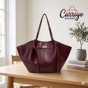 May include: A burgundy leather tote bag with long handles, a rectangular shape, and a gold-colored clasp. The bag is sitting on a wooden table. The brand name "Carriyo Designs" is visible in the upper right corner.