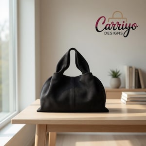 Peut inclure: Un sac à main noir avec une forme arrondie et des anses souples drapées est posé sur une table en bois clair. Le sac est le point central, avec un design minimaliste. La marque "Carriyo Designs" est visible en arrière-plan.