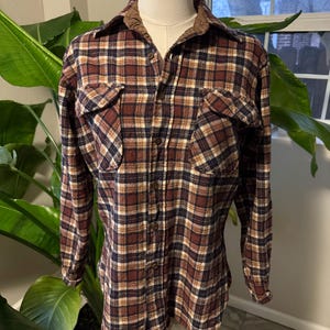 May include: A long-sleeved plaid shirt in shades of brown, burgundy, navy, and cream. The shirt features two chest pockets and a brown corduroy collar. The button-down shirt is displayed on a mannequin.