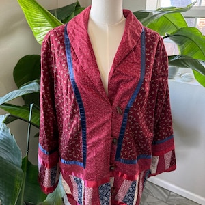 May include: A burgundy jacket with a patchwork design. The jacket features a notched collar, long sleeves, and wooden buttons. Blue ribbon accents the front and sleeves. The bottom hem has a patchwork design with red ribbon trim. The jacket is displayed on a mannequin.