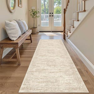 May include: A rectangular, beige runner rug with a distressed, vintage-inspired design. The rug features a subtle border and is placed on a hardwood floor. The runner extends towards a set of double doors and a staircase.