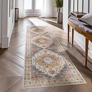 May include: A rectangular runner rug with an intricate, symmetrical pattern in shades of beige, gray, and rust. The rug features a central medallion design and a border with geometric motifs. It is placed on a wooden floor in a hallway.