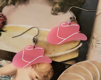 May include: A pair of pink cowboy hat earrings with white trim and silver-colored ear wires. The earrings are displayed on a vintage-style background with a soft focus.