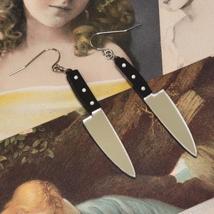 May include: A pair of novelty earrings shaped like miniature knives. Each earring features a black handle with three silver studs and a silver blade. The earrings hang from silver fish hook ear wires. The earrings are displayed on a vintage-style background.