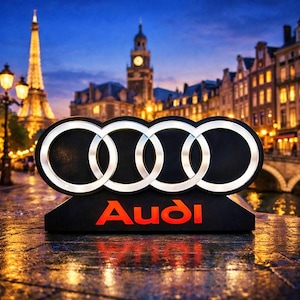 May include: A black Audi logo with silver rings and red lettering, set against a blurred cityscape at dusk. The iconic four rings are prominently displayed above the brand name.