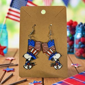 May include: A pair of patriotic earrings featuring a cartoon dog wearing a Uncle Sam hat and holding a flag. The earrings are attached to a brown card with a hole at the top. The background includes American flags and decorative stars.