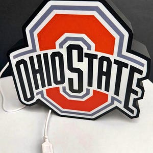 May include: A black, orange, white, and gray Ohio State logo light with the text "OHIO STATE". The light has a USB cable attached.