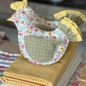 May include: Two handmade chicken-shaped oven mitts with floral fabric, yellow accents, and green polka dot details, resting on a stack of yellow and white kitchen towels. The mitts have a pocket and ruffled edges.