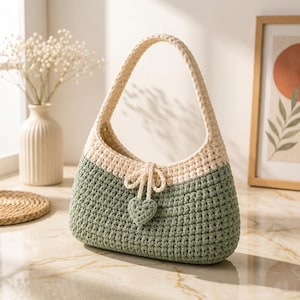 May include: A two-toned crochet handbag featuring a sage green base and a cream-colored upper section with a single handle. The bag includes a decorative drawstring closure finished with a small, matching sage green crocheted heart charm.