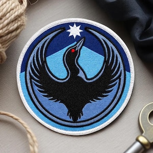 May include: Embroidered patch featuring a stylized black bird with outstretched wings, a red eye, and a white star. The patch has a circular design with blue and white accents, and a white border. The patch is likely for clothing or accessories.