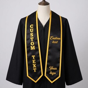 May include: Black graduation gown with a gold-trimmed sash. The sash has the words "CUSTOM TEXT" embroidered vertically and "Custom text" and "Your logo" embroidered horizontally in gold. The gown is displayed on a mannequin.