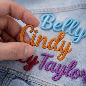 May include: Embroidered name patches in orange, red, purple, and blue script lettering. The patches are shown on a denim jacket. The name "Cindy" is being held up by a hand. Other names include "Belly" and "Taylor".