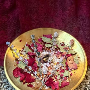 May include: A gold-colored bowl filled with dried rose petals, bay leaves, and small crystals. Three silver wire stems are adorned with various gemstones. The background is a deep red fabric.