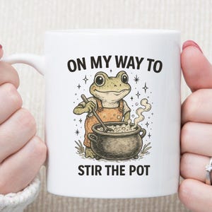 May include: White ceramic mug featuring a cartoon frog in an orange apron stirring a pot. The mug has the text "ON MY WAY TO STIR THE POT" in black. The frog is green and the pot is dark brown.