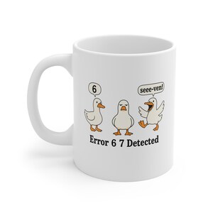 Op de afbeelding: Witte keramische mok met een cartoon illustratie van drie eenden. Eén eend zegt "6", een andere kijkt chagrijnig en de derde roept "seee-ven!". De tekst "Error 6 7 Detected" staat onder de eenden.