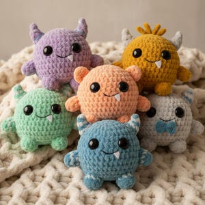 May include: A collection of six crocheted monster toys in various colors, including purple, orange, green, yellow, blue, and gray. Each monster has large black eyes, a single tooth, and small horns or ears. The toys are arranged on a soft, textured surface.