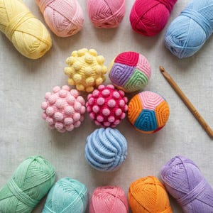 Puede incluir: Una colección de bolas de ganchillo y ovillos de lana de colores en varios tonos de rosa, amarillo, azul, verde y naranja. También hay un ganchillo de madera. Las bolas tienen superficies texturizadas y la lana está bien organizada.