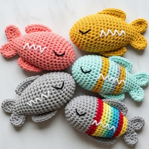 May include: Five crocheted fish toys in various colors: pink, yellow, blue, gray, and a rainbow pattern. Each fish has a white zig-zag mouth and a black curved eye, creating a whimsical design. These handmade toys are soft and cuddly.