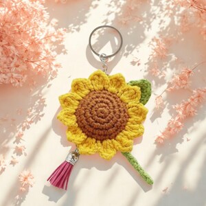 May include: A handmade crochet sunflower keychain. The sunflower has yellow petals and a brown center, with a green leaf and stem. A pink tassel hangs from the bottom. The keychain ring is silver.