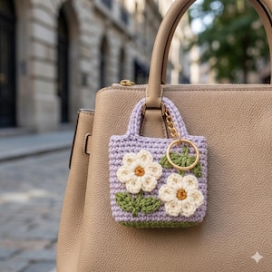 May include: A small, lavender crochet bag charm with two cream-colored daisy appliqués and green leaves. The charm is attached to a gold-colored keyring and hangs from the handle of a beige handbag.