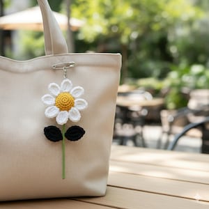 May include: A cream-colored tote bag with a crocheted daisy charm. The daisy has white petals, a yellow center, black leaves, and a green stem. The charm is attached with a safety pin.