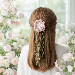 Op de afbeelding: Een gehaakte haaraccessoire met een roze bloem en groene bladeren. De bloem is met een vlecht aan het haar bevestigd. Het accessoire is handgemaakt en voegt een vleugje elegantie toe, geïnspireerd door de natuur, aan elke haarstijl.
