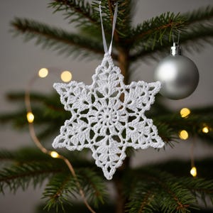 Puede incluir: Adorno navideño de copo de nieve blanco tejido a ganchillo, colgado de un árbol de Navidad. El copo de nieve tiene un diseño calado detallado y está suspendido por una cinta blanca. También se ven una bola plateada y ramas de árbol.