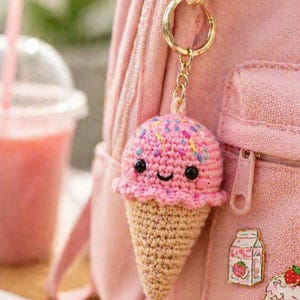 May include: A handmade pink crocheted ice cream cone keyring with a smiling face and colourful sprinkles. The ice cream cone is attached to a gold-coloured keyring and is hanging on a pink backpack.
