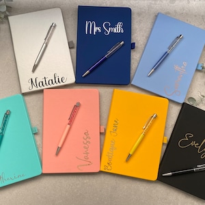 May include: A collection of eight personalised notebooks with matching pens. The notebooks are in various colours, including silver, blue, light blue, pink, yellow, and black. Each notebook has a name or title written in gold lettering, such as "Natalie", "Mrs. Smith", "Evelyn", and "Boutique Jane".