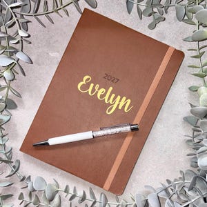 May include: A brown notebook with a vertical elastic band closure, featuring the name Evelyn and the year 2027 printed in gold on the front cover. A white pen with a clear, crystal-filled barrel is placed horizontally across the notebook cover.