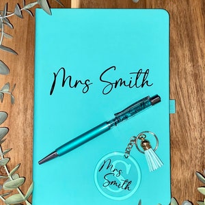 May include: A turquoise notebook with the name "Mrs Smith" written on the front. A turquoise pen with glitter and a keyring with a turquoise circle that says "Mrs Smith" are on top of the notebook.