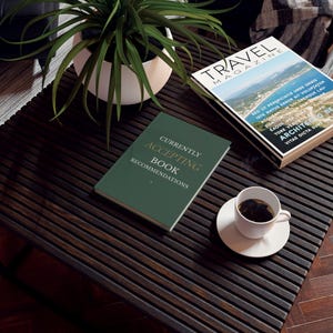 May include: A dark green hardback book with the text "CURRENTLY ACCEPTING BOOK RECOMMENDATIONS" in gold lettering, placed on a dark slatted table next to a white coffee cup, a travel magazine, and a potted green plant.