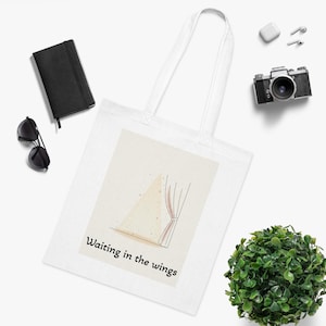 May include: White tote bag with a graphic of a stage curtain and the text "Waiting in the wings." The bag is surrounded by a black notebook, sunglasses, a camera, earbuds, and a small green plant.