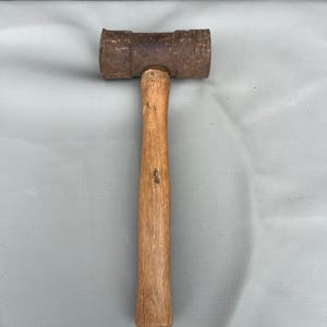 May include: A vintage mallet with a wooden handle and a rectangular, rust-colored head. The handle is a light brown color and shows signs of wear. The tool is designed for striking or shaping materials.