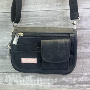 May include: Black crossbody bag with a black adjustable strap. The bag has a front pocket and a zippered top. The brand name "Mudd" is displayed on a pink rectangular label. The bag is decorated with a subtle floral and heart pattern.