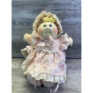 May include: A vintage Cabbage Patch Kid doll wearing a pink and white striped dress with floral accents and a matching bonnet. The doll has a round face, blue eyes, and blonde curly hair. The dress features lace trim and a ribbon bow.