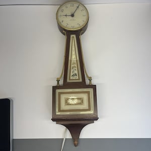 May include: A wall-mounted wooden clock with a circular face featuring black numerals and hands. The clock body has a dark brown finish with gold-toned metal accents, a central vertical panel with floral detailing, and a rectangular lower section.