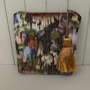 May include: A rectangular cosmetic bag with a colorful abstract pattern in shades of brown, green, blue, and red. The bag has a gold-colored tassel attached to the zipper.
