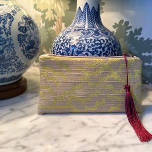 May include: A rectangular zippered pouch featuring a textured beige fabric base with a vibrant neon yellow zigzag pattern. A deep burgundy tassel is attached to the silver-toned zipper pull, providing a distinct contrast to the light-colored textile.