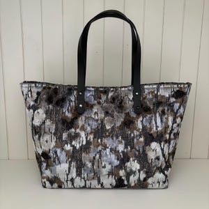 May include: A large tote bag with a textured pattern in shades of gray, brown, and black. The bag has two black leather handles attached with silver rivets. The bag is designed to be carried over the shoulder.