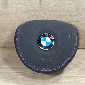 Könnte beinhalten: Ein schwarzer Airbag für das Lenkrad mit dem BMW-Logo in der Mitte. Der Airbag hat rote, weiße und blaue Nähte am Rand und das Wort "AIRBAG" ist unten aufgedruckt.