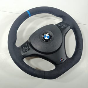 Könnte beinhalten: Ein schwarzes Lenkrad mit blauen Nähten und einem blauen Streifen oben. In der Mitte befindet sich ein BMW-Logo und unten rechts ein M-Logo. Das Lenkrad hat Knöpfe und einen strukturierten Griff.