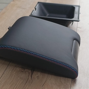 Może przedstawiać: Czarny podłokietnik samochodowy z czerwonymi i niebieskimi szwami. Podłokietnik jest prostokątny z zakrzywionym wierzchem i małą, dołączoną przegrodą. Widoczny jest oddzielny, czarny plastikowy pojemnik do przechowywania.